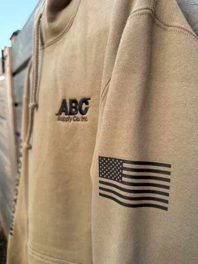 Tan hoodie with a left-chest ABC Supply logo and a black flag printed on the sleeve